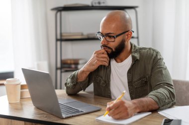 Laptop ekranına bakan, not defterine yazan, iş yerinde çalışan Latin bir adam. Konsantre erkek notlar alıyor, web sitesinde ders çalışıyor, boş alan