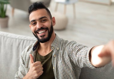 Mutlu genç Arap çocuk selfie çekiyor ve parmağını kaldırıyor, cep telefonu kamerasına gülümsüyor, evdeki kanepede oturuyor. Pozitif Orta Doğulu bir adamın içeride kendi fotoğrafını çektiği portresi.