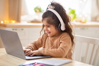 Kablosuz kulaklıklı sevimli küçük kız evde dizüstü bilgisayar kullanıyor, klavyesini daktilo ediyor, ev ödevlerini çevrimiçi yapıyor, uzaktan öğrenmenin ya da internette oyun oynamanın tadını çıkarıyor.