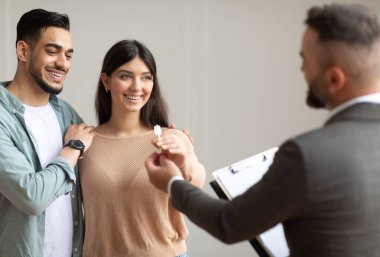 Mutlu gülümseyen Orta Doğulu çift gelecekteki yeni dairenin anahtarını alıyor. Takım elbiseli profesyonel bir emlakçı genç alıcılara anahtar veriyor. İpotek, Satılık Mülkiyet