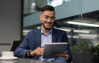 Güzel takım elbiseli neşeli Arap milenyum adamı masada oturuyor, dijital tablet kullanıyor, yeni iş yeri uygulamalarını kontrol ediyor ve gülümsüyor, kopyalama alanı kullanıyor. Modern teknolojiler ve iş dünyası