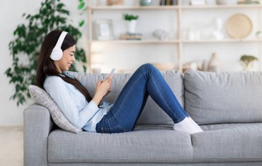 Asyalı bir kızın akıllı telefonuyla koltuğa uzanması, kulaklıkla müzik dinlemesi, evde boş zaman geçirmesi, kenar manzaranın tadını çıkarması.