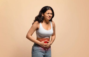 Hasta genç Hintli kadın mide ağrısı çekiyor, karnına kırmızı iltihaplı bölge ile dokunuyor, doğulu kadın regl ağrısı çekiyor ya da gastrointestinal hastalık geçiriyor, bej arka planda poz veriyor.