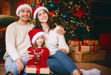 Üç kişilik genç bir aile Noel ağacı üzerinde poz veriyor, elinde Noel hediyesi tutan bir kız çocuğu.