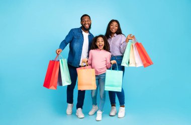 Alışveriş eğlencesi. Afro-Amerikalı neşeli adamın vücut portresi, bayan ve heyecanlı genç kız kağıt torbalarıyla poz veriyor, mavi stüdyoda yan yana duruyor, satışların ve alışverişlerin keyfini çıkarıyor.
