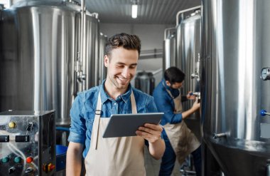 Aile işi, bira fabrikasında çalışan bira üreticileri, kontrol üretimi. Önlüklü gülümseyen bin yıllık yakışıklı adam tablet kullanıyor, verileri el yapımı içecekle kontrol ediyor, boş alan var.