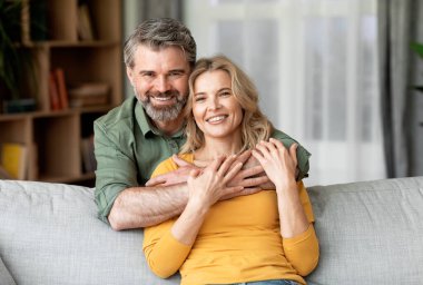 Portrait Of Smiling Middle Aged Spouses Posing In Home Interior, Romantic Mature Couple Embracing And Looking At Camera While Resting On Sofa In Cozy Living Room, Enjoying Time Together, Free Space
