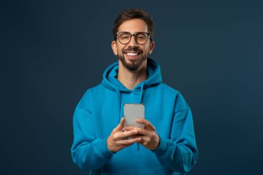Elinde akıllı bir telefonla gülümseyen genç adam portresi kapüşonlu ve gözlüklü adam mavi stüdyonun arkasında dururken cep telefonu kullanıyor.