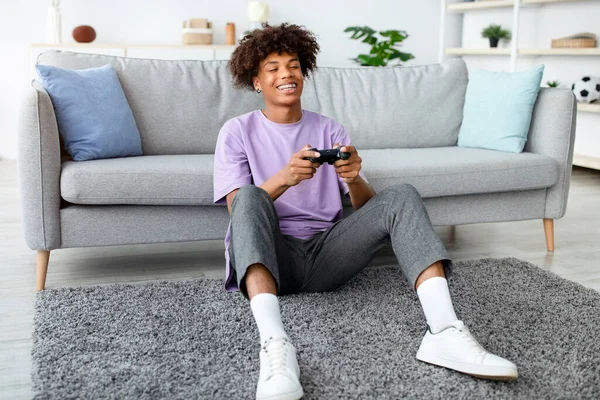 Playstation 'da video oyunundan hoşlanan mutlu siyahi genç, evde uzun süre eğleniyor. Joy Afro-Amerikan ergen oyuncu elinde joystickle yerde oturmuş bilgisayar oyunu oynuyor.