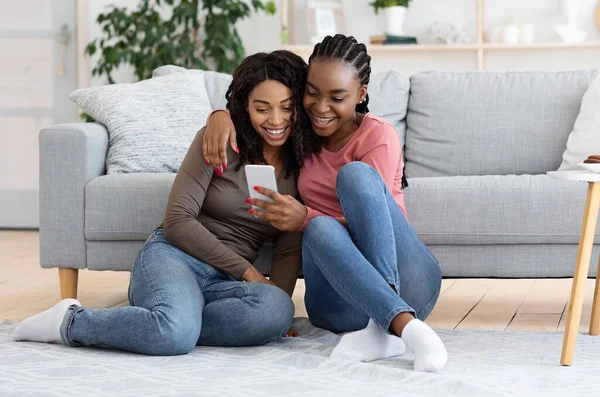 İki neşeli Afro-Amerikalı kız kardeş sosyal medyayı akıllı telefondan kontrol ediyor, oturma odasında yerde oturuyor. Mutlu siyahi bayanlar evde dinlenirken yeni mobil uygulama kullanıyorlar, boş alan.
