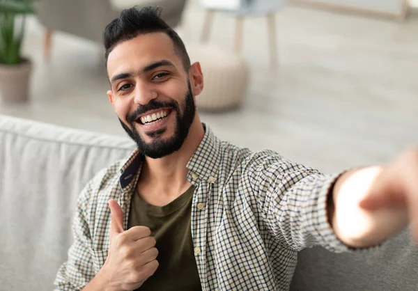 Mutlu genç Arap çocuk selfie çekiyor ve parmağını kaldırıyor, cep telefonu kamerasına gülümsüyor, evdeki kanepede oturuyor. Pozitif Orta Doğulu bir adamın içeride kendi fotoğrafını çektiği portresi.