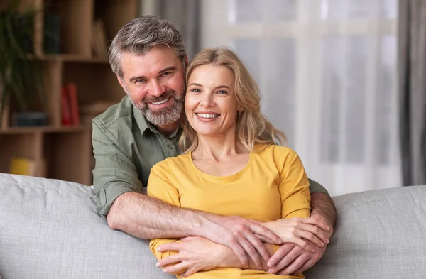 Orta yaşlı mutlu bir koca ve ev içinde poz veren, eşleri seven, oturma odasında koltukta dinlenirken kameraya gülümseyen, arkadan sarılan bir erkek.