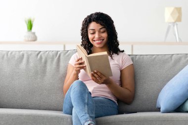 Evde eğitim, tek başına eğitim, not defteriyle planlama günü ve boş zamanlarında akıllı bir hobi. Tatmin olmuş inek, Afro-Amerikalı bir bayan minimalist oturma odasında, boş bir odada koltukta kitap okuyor.