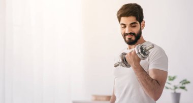 Kapalı alan 19, aktif hobi ve boş zaman, evde güç eğitimi, spor. Gülümseyen Arap sakallı genç adam modern basit oturma odasında dambılları kaldırıyor penceresi olan, kopyalama alanı olan