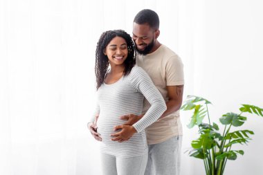 Binlerce yıllık gülümseyen Afro-Amerikan erkeği hamile kadınına sarılıp karnına dokunduğu için mutlu. Minimalist beyaz yatak odası iç mekanı, boş alan. Evde mutlu bir ilişki, aile planlaması.