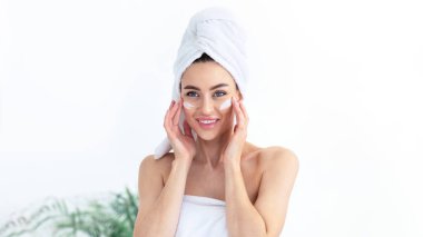 Mükemmel, sağlıklı, parlak cilt, yaşlanma önleyici bakım ve evde spa günü. Havluya sarılmış, yüzüne krem süren, masaj yapan ve aynaya bakan 30 'lu yaşlardaki güzel kadın.