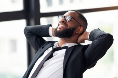 Gülümseyen, gözlüklü rahat siyah iş adamının portresi sandalyeye yaslanıyor, el ele tutuşuyor, işten sonra modern ofiste dinleniyor. Takım elbiseli adam günün sonunda eğleniyor.