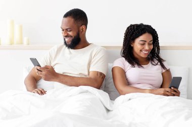 Glad young black female ignores her husband sitting on bed and look at phone in bedroom interior, copy space. Treason, addiction on social networks and technology, personal information and new normal