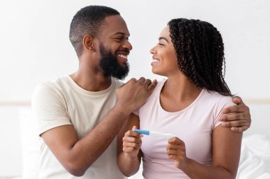 Satisfied smiling millennial african american man look at cheerful cute woman hold positive pregnancy test, celebrate great news in bedroom interior. Positive emotions, family planning, parenting