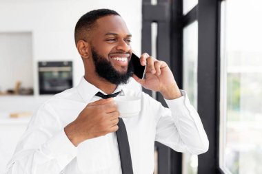 Kurumsal İletişim. Beyaz gömlek ve kravat takmış gülümseyen Afrikalı Amerikalı bir adamın portresi. Sabah cep telefonuyla konuşuyor. Elinde bir fincan sıcak kahveyle pencerenin önünde duruyor.