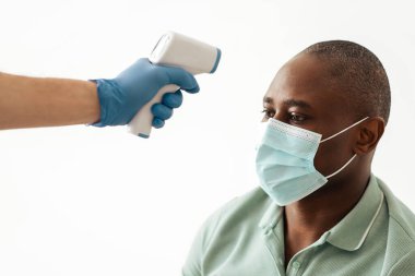 Covid-19 virüsünü önleyin ve nüfus aşısı için hazırlanın. Koruyucu maskeli hemşire enjeksiyondan önce yetişkin Afrikalı Amerikalı hastalarda kızılötesi termometreyle ısıyı ölçer.