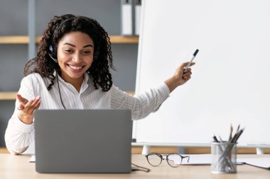 Video konferansı, e-öğrenme, çevrimiçi eğitim COVID-19 virüsünden sosyal uzaklık koruması. Mutlu siyah kadın öğretmen tahtayı işaret ediyor, kuralları açıklıyor, evdeki laptop web kamerasına bakıyor.