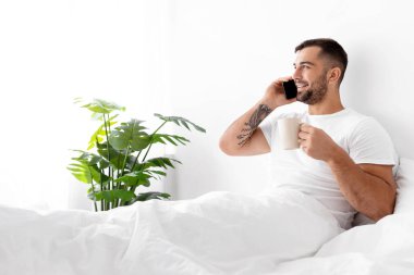 Kafkas kaslı, mutlu genç adam beyaz yatakta oturur, kahvesini içer ve yatak odasının içinde telefonla konuşur. Evde dinlenmek ve dinlenmek, adam iletişimi seviyor, günaydın, boş zaman ve tatil.