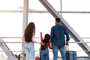 Hava Yolculuğu. Kızı havaalanında uçaktan ayrılan bir kıza bakan aile, küçük bir kız çocuğu, Terminalde elinde bavullarla bekleyen genç ebeveynlerin ve kız çocuğunun arka plan görüntüsü.