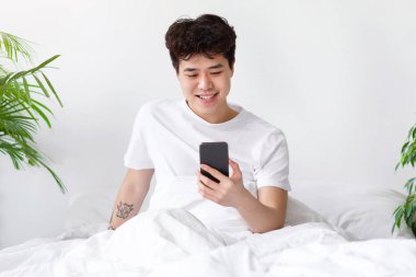 Harika mesaj, online teklif, iyi haber, sabahları pozitif ifade. Gülümseyen Asyalı genç, beyaz tişörtlü genç adam akıllı telefona bakıyor ve yatak odasında bitkilerle oturuyor.