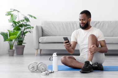 Beyaz spor giyimli, sakallı, telefonda yazan gülümseyen siyah bir adam oturma odasının içinde yerde oturuyor. Spor malzemeleri, eğitim uygulaması ve karantina süresi dolan vücut bakımı