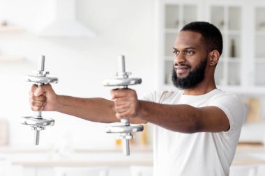 Gülümseyen siyah sakallı, beyaz spor giysili genç bir sporcu Covid-19 sırasında minimalist mutfak iç kısmında halterlerle el egzersizi yapıyor. Kaslı ve güçlü adam evde vücut çalışıyor.