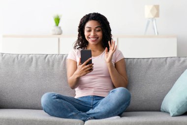 Covid-19 tecrit, çevrimiçi arama, modern teknolojiyle görüşme ve sohbet. Bin yıllık güzel Afro-Amerikan kadınının akıllı telefona bakması ve el sallayarak oturma odasının iç tarafındaki webcam 'e el sallaması çok güzel.