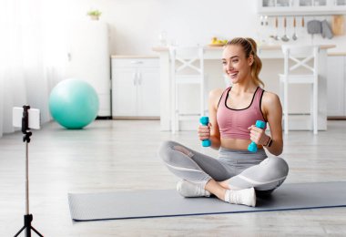 Formda blogcu, ders, online ders ve modern teknoloji için egzersiz videosu yapıyor. Neşeli milenyum güzel kadın spor giyimli, halterli halterde oturuyor. Evdeki internet kameralarına bakıyor.