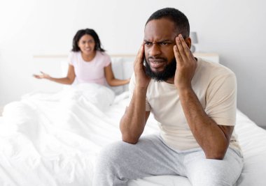 Unhappy young black wife freaks out and yells at her sad husband, man suffers from erectile dysfunction and headache, sits on bed in bedroom interior. Impotence, quarrel, scandal, family problems