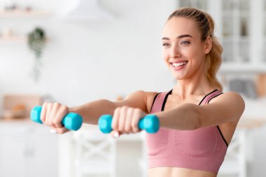 Neşeli, mutlu, beyaz, güzel, sarışın kadın evde dambılları kaldırıyor, kapalı alanda çalışıyor ve kollar için egzersiz yapıyor, boş alan. Sosyal mesafe ve eğitim, covid-19 salgını ve yeni normal