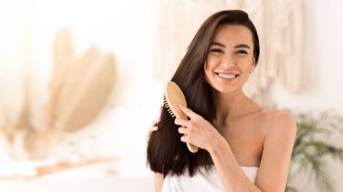 Kadınlar boş vakitlerinde evde saç bakımını, tedaviyi ve spa gününü yaparlar. Havluya sarılmış, uzun ve sağlıklı saçlarını tarak ile tarayan mutlu, genç, tatlı esmer kadın.