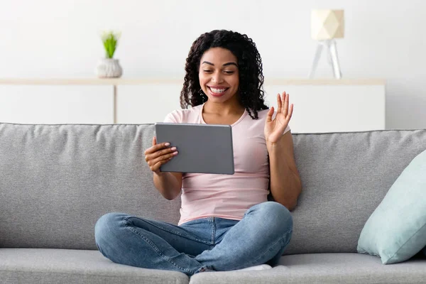 Covid-19 karantinası sırasında çevrimiçi toplantı, evde çalışmak ve sosyal mesafeler, teknoloji. Tatmin olmuş Afro-Amerikan kadın tablet webcam 'e bakıyor ve el sallıyor.