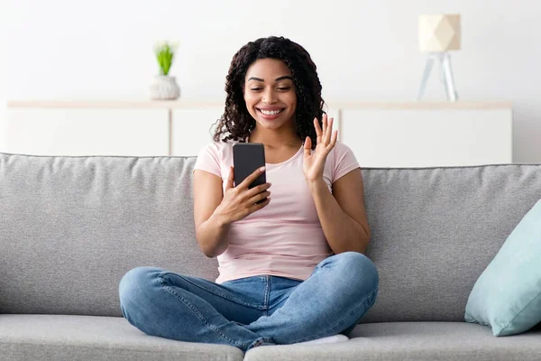 Covid-19 tecrit, çevrimiçi arama, modern teknolojiyle görüşme ve sohbet. Bin yıllık güzel Afro-Amerikan kadınının akıllı telefona bakması ve el sallayarak oturma odasının iç tarafındaki webcam 'e el sallaması çok güzel.