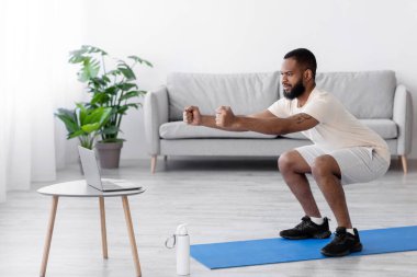 Konsantre olmuş, siyah sakallı, beyaz spor elbiseli bir sporcu oturma odasının içinde, profilde. Fitness günlüğü, sosyal uzaklık, aygıt, covid-19 sırasında yeni normal ve evde çalışma