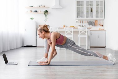 Evde Covid-19 karantina, esneme, modern cihazlarla yoga ve çevrimiçi eğitmen. Neşeli, genç, çekici, sarışın kadın spor giyimli minimalist mutfak iç mekanında bacaklar için egzersiz yapıyor.