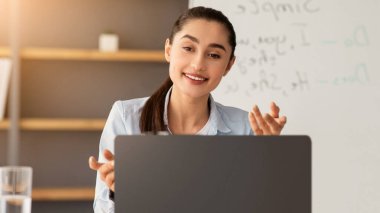 Çevrimiçi ders ve eğitim, yeni normal, covid-19, yakınlaştırma dersi ve sosyal mesafe. Gülümseyen Arap bayan öğrencilerle konuşur, oturma odasındaki akıllı telefona bakar, dizüstü bilgisayarla el kol hareketi yapar.