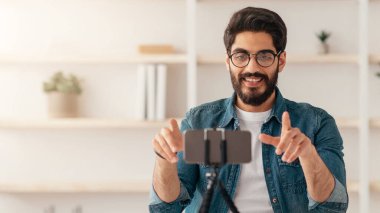 Evden yayın yapan, akıllı telefon ve tripodu kullanan, oturma odasında oturan ve kopyalayan olumlu Arap erkek etkili. Gülümseyen adam video kaydedicisi cep telefonu kamerasına konuşuyor