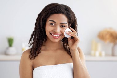 Cilt bakımı, makyaj giderici, temizlik ve evdeki rutin prosedürler. Gülümseyen bin yıllık Afro-Amerikan kadın beyaz havluyla yüzünü pamuk süngerle siliyor. Yatak odasının içinde, boş bir yerde.