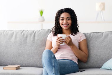 Hobiler, okuma ve boş zaman, tatilde evde taze kahvenin tadını çıkar. Neşeli bin yıllık güzel Afro-Amerikan kadın oturma odasının içinde en sevdiği sıcak içeceği içiyor.