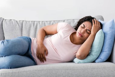 İyi hissetmiyor, regl, grip, migren ve hastalık geçiriyor. Üzgün, genç, Afro-Amerikan kadın, kanepede uzanmış, baş ağrısı ve oturma odasındaki karın ağrısından muzdarip.