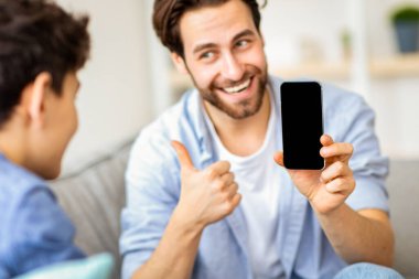 Bu uygulama gibi. Mutlu genç baba oğluna boş siyah ekranlı akıllı telefonu gösteriyor ve baş parmağını gösteriyor, maket için uygun bir yer. Modern teknolojiler, yeni uygulama kavramı