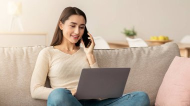 Ev ofisi, müşteriyle çalışmak, evdeki iş kadını. Gülümseyen genç Arap kadın müşteriyle telefonda konuşuyor ve dizüstü bilgisayara bakıyor, oturma odasındaki kanepede oturuyor.