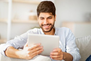 Teknoloji konsepti. Modern dairesindeki koltukta dijital tablet kullanan mutlu bir Arap 'ın portresi. Heyecanlı adam haberleri okuyor, internette geziniyor, oturma odasındaki kanepede dinleniyor.