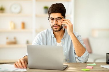 Uzaktan kumanda. Yoğunlaşmış Arap erkek serbest çalışan cep telefonuyla konuşuyor ve merkez ofiste dizüstü bilgisayarla çalışıyor. Adam işyerinde oturuyor, bilgisayar kullanıyor ve telefon görüşmesi yapıyor.
