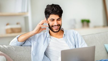 Müşteri desteği. Neşeli Arap adam, bilgisayarını kullanarak çağrı merkezi operatörüyle telefonda konuşuyor. Bilgisayarda çalışan serbest çalışan, oturma odasındaki kanepede oturan